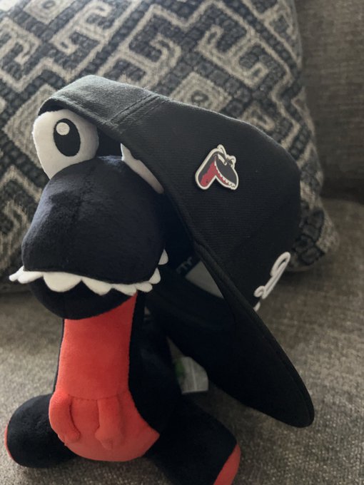 A plush toy wearing a black baseball cap with a dinosaur logo, sitting on a gray patterned cushion.