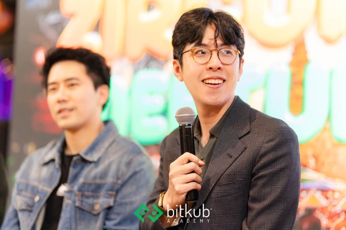 Two men at an event with a 'bitkub ACADEMY' logo visible; the man in the foreground wears glasses and a dark blazer while speaking into a microphone, with another man in a denim jacket standing behind him.