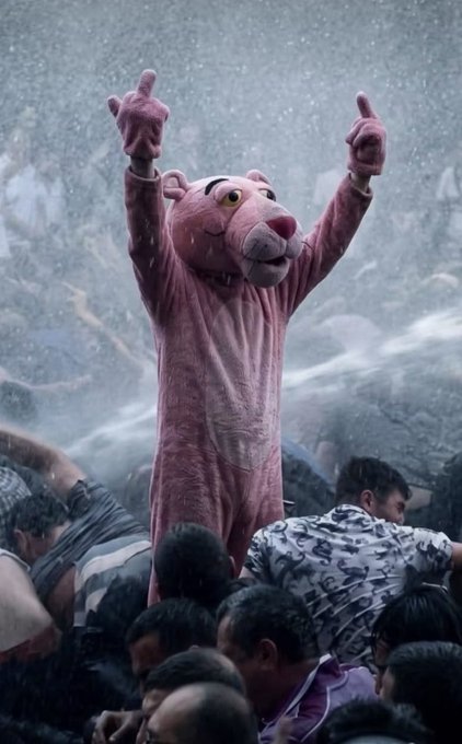 A person in a pink panther costume with both arms raised triumphantly above a crowd of people in a rainy setting.