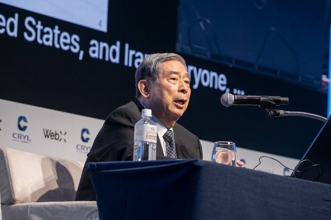 A distinguished older man, presumably SBI Chairman Norio Kato, speaks into a microphone at the WebX 2026 conference, with sponsor logos for "CRYL" and "WebX" visible on the backdrop behind him. The setting aligns with the post's context of a keynote speech about tokenization and real-world asset (RWA) scaling.
