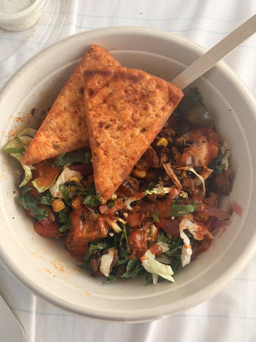 A white bowl contains a mixed dish with corn chips, greens, and a reddish sauce, with a compostable fork resting inside.
