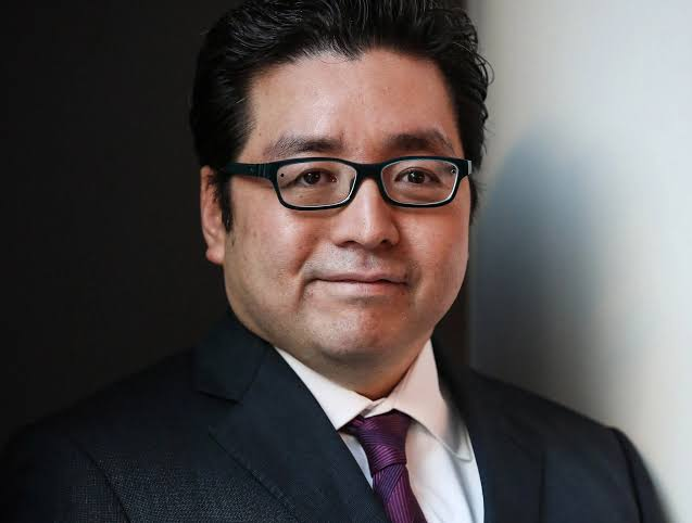The image features a man wearing glasses and a suit standing against a white wall. The man has short dark hair and is smiling at the camera. There are no texts or other objects present in the image, so we can only focus on the man's attire and facial expression.