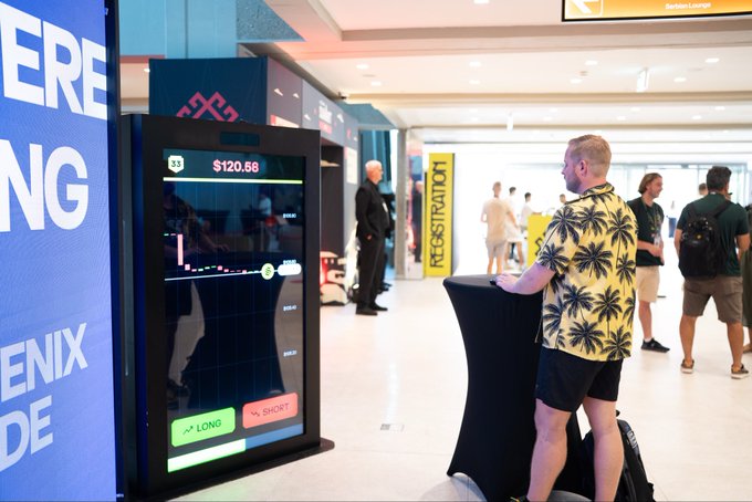 A photo of a man in a yellow tropical shirt standing at a digital kiosk trading interface displaying $120.68 with LONG/SHORT buttons at what appears to be a crypto conference.