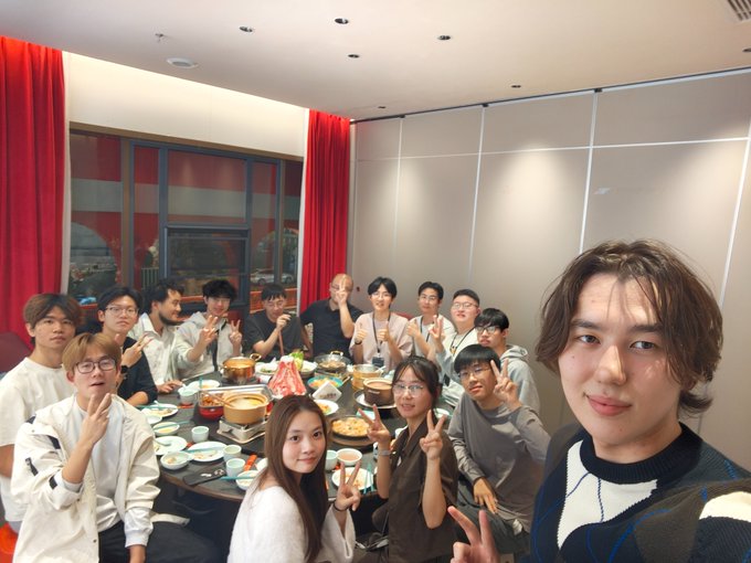 A group selfie of about fourteen young people gathered around a large dining table filled with various dishes, all smiling and making peace signs at the camera.