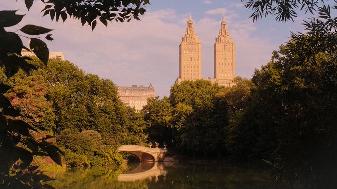 A scenic view of a stone bridge over a calm pond, framed by trees, with two prominent twin towers (the San Remo building) rising in the distance against a partly cloudy sky.