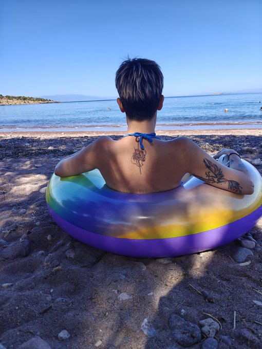 A young boy with temporary tattoos on his back sits in a colorful inflatable ring on a rocky beach, facing the calm ocean.