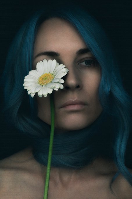 A close-up portrait of a woman with blue hair holding a white daisy over one eye, creating a striking visual contrast against a dark background.