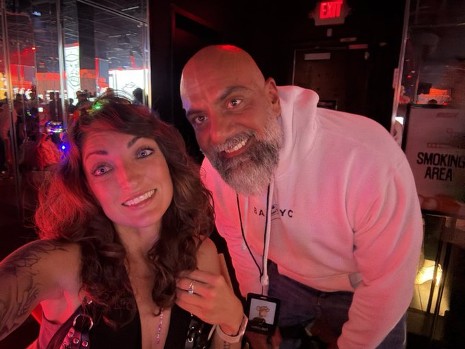 A selfie of a smiling woman and a bearded bald man in a white "NYC" hoodie, standing together in a dimly lit venue near a door marked "SMOKING AREA."