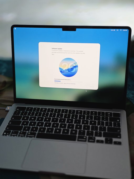 A MacBook laptop displaying a software update screen for macOS version 26 with the 'Downloading macOS Tahoe' message.