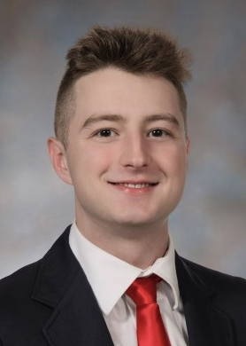 A young man with a bright smile poses for a professional portrait, wearing a navy suit, white shirt, and red tie, suggesting he is being recognized for an achievement. The post hints that he is among students advancing to the state-level competition at Penn, likely reflecting his academic or extracurricular success.