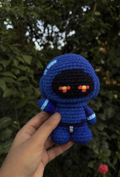 A hand holding a small crocheted amigurumi figure of a blue robot with glowing red eyes, positioned outdoors with green foliage in the background.