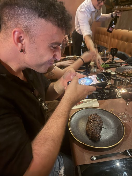 A man smiles while photographing a grilled steak at a restaurant, capturing the meal on his phone to share with his crypto newsletter collaboration "La Entremesa."