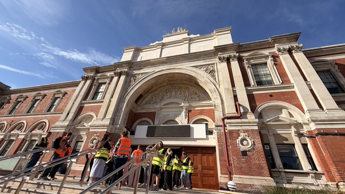 A group of people wearing high-visibility vests gather on the steps in front of an ornate, historic brick building with elaborate white stone carvings, columns, and an arched entrance.