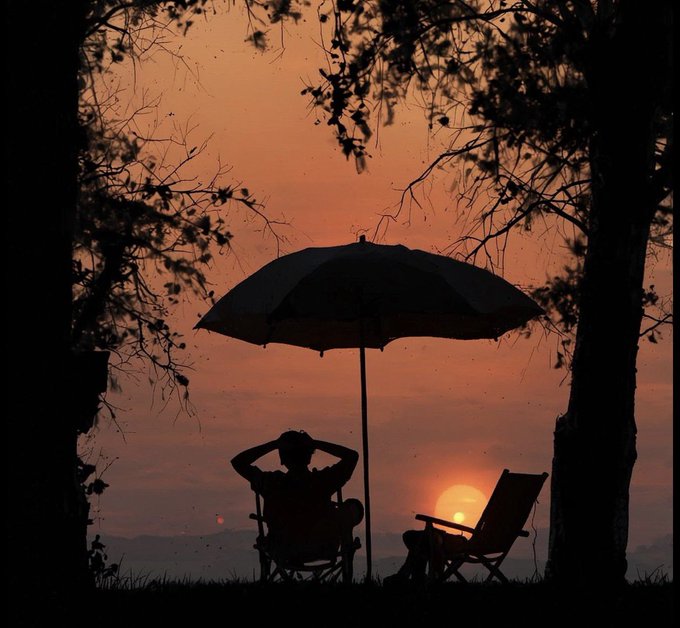 A silhouetted figure relaxes under an umbrella beside an empty chair, watching a glowing sunset framed by tree branches. The serene scene evokes the peaceful, escape-like feeling suggested by the accompanying "GN CT @RiverdotInc @River4fun $river" post, hinting at tranquility by the water.