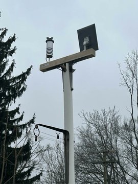 A photograph shows a utility pole supporting a small solar panel and what appears to be an owl decoy or weather sensor, with bare trees visible in the background.