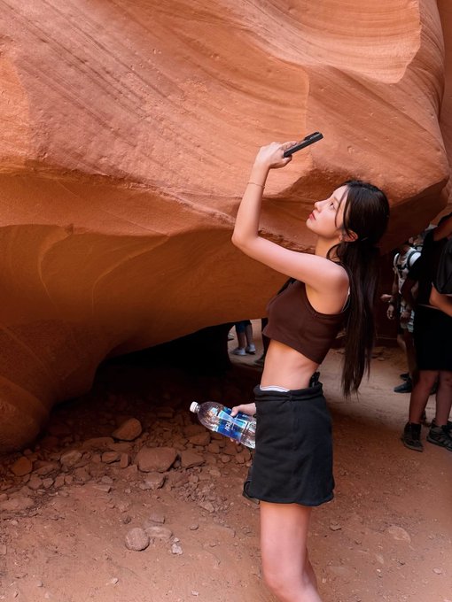 A young woman in a brown crop top and black skirt takes a photo with her phone while holding a water bottle, standing against a large orange rock formation.