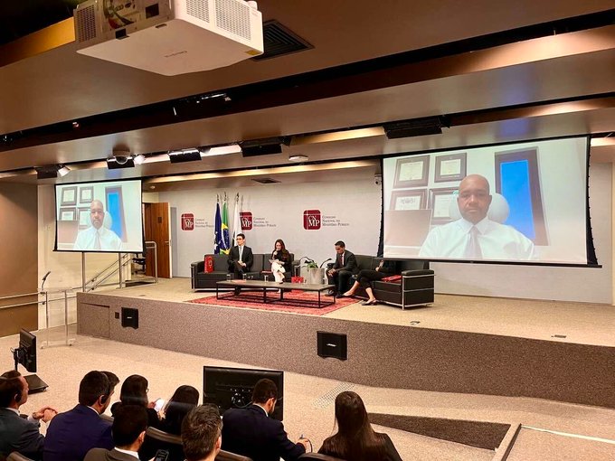 A panel discussion on digital asset recovery hosted by Brazil's National Council of the Public Ministry (CNMP) and DCGG, featuring John Hurston (General Counsel at TRON) appearing via video call on screens as in-person panelists seated on stage discuss consumer protections and fund restitution before a seated audience.