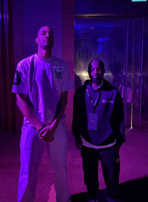 Two men standing in a room with purple lighting, one wearing a white t-shirt and sunglasses, the other in a black jacket with a lanyard.