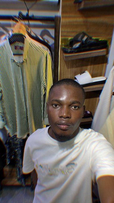 A man in a white t-shirt takes a selfie in what appears to be a closet, with striped shirts hanging behind him and a distorted, colorful visual effect applied to the image.