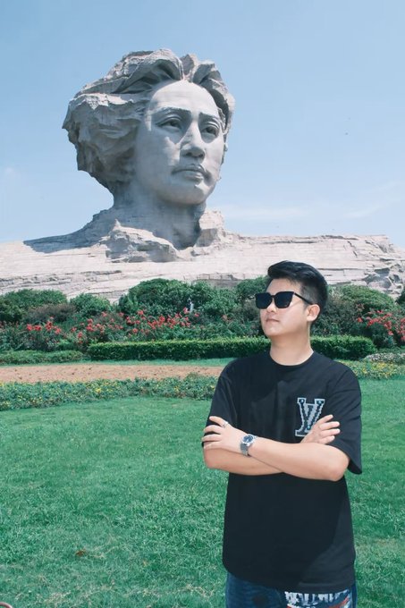 A young man in a black "V" shirt and sunglasses poses confidently in front of a giant stone bust of Mao Zedong at Orange Isle (橘子洲) in Changsha, with the post's "国庆快乐呀" (Happy National Day) greeting tying the scenic visit to China's National Day celebrations.