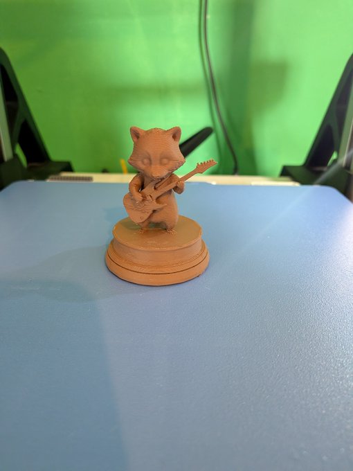 A 3D-printed figurine of a small, stylized bear-like creature playing a guitar, sitting on a circular base atop a blue 3D printer bed.