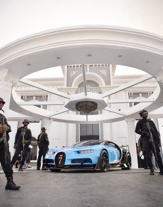 A blue and black Bugatti Chiron Pur Sport supercar is parked under the circular porte-cochère of a grand white building, surrounded by security personnel in tactical gear.