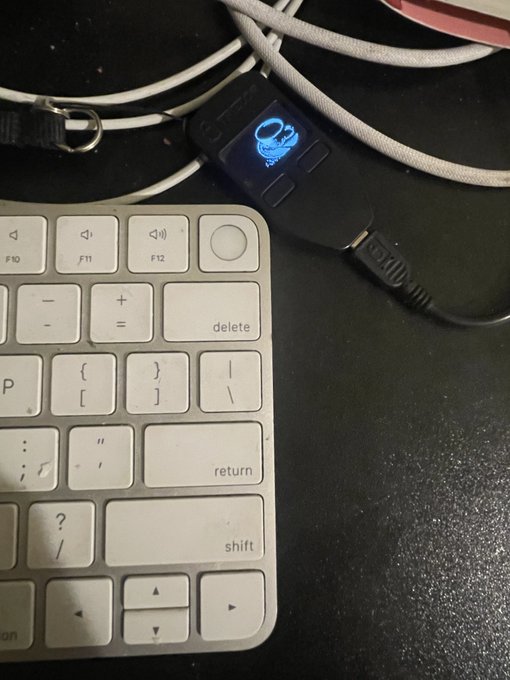 A close-up of a small blue LCD display device showing a coffee cup icon, connected via USB cable next to a white Apple Magic Keyboard on a black desk.