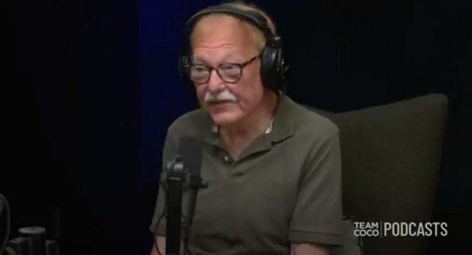 An older man with glasses and headphones speaking into a microphone during a 'Team Coco' podcast recording.