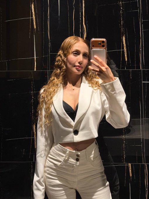 A woman with long curly blonde hair wearing a white blazer and white skirt taking a mirror selfie with her phone.