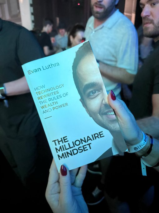 A hand with red nail polish holding up the book 'The Millionaire Mindset' by Evan Luthra, subtitled 'How Technology Rewrites the Rules of Wealth and Power,' at what appears to be a networking event.