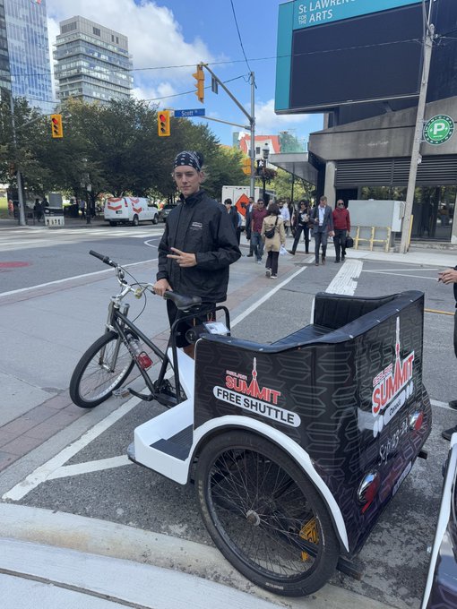 A person wearing a black jacket and beanie stands next to a pedicab labeled "START SUMMIT FREE SHUTTLE" on a city street corner near a sign for "St. Lawrence for the ARTS".