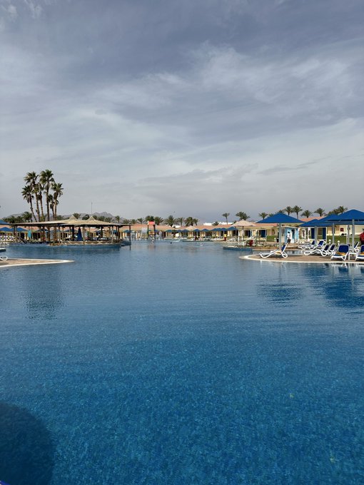 A serene resort pool scene with palm trees, cabanas, and lounge chairs under a partly cloudy sky, capturing the tranquil "Good Morning" vibe of a vacation day just beginning.