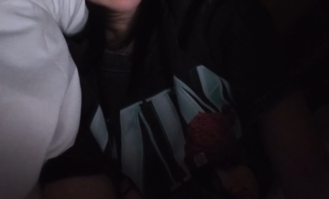 A dimly lit close-up photo captures a person wearing a black shirt featuring large white "MINA" lettering, with a small red plush toy tucked inside it, paired with the caption "THE MINA SHIRT???" to express excitement over the fan merchandise.