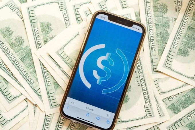 A smartphone displaying the USDC stablecoin logo rests on a fan of US hundred-dollar bills.