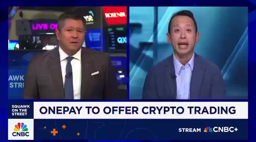 The image shows two men standing next to each other, with a news report about Bitcoin trading taking place on a television screen behind them. The TV show is discussing the benefits of Bitcoin trading for its one-billion customers. One man is wearing a suit while the other has a tie, and they are both looking at the camera. They appear to be experts in the field, possibly discussing the advantages or disadvantages of using Bitcoin as a payment method.