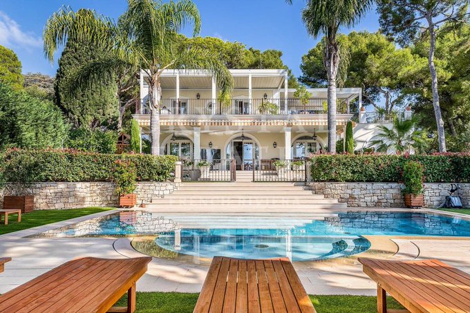 Luxury Mediterranean-style two-story villa with a large swimming pool, sun loungers on a manicured lawn, lush palm trees, and outdoor patio furniture under a clear blue sky.