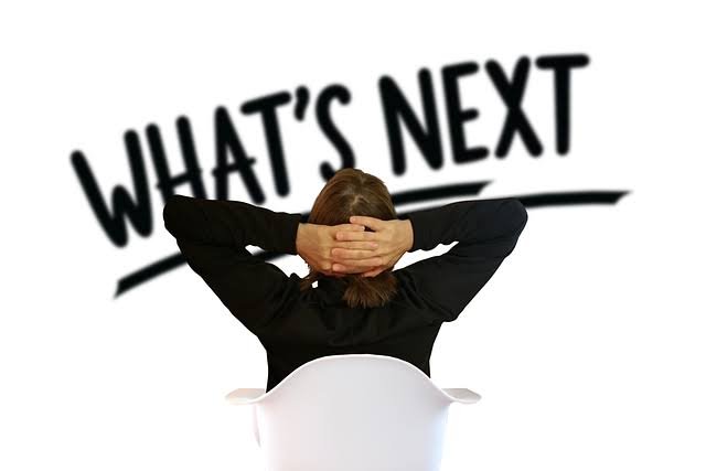 A woman in black business attire sits in a white chair with her hands clasped behind her head, viewed from behind, with the bold black text "WHAT'S NEXT" displayed above her on a white background.