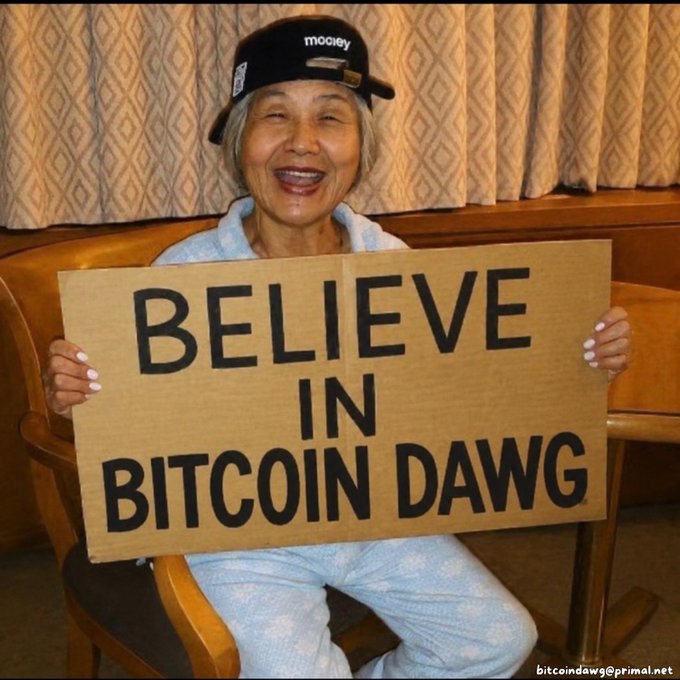 An older woman wearing a black 'mooney' cap and a light blue robe smiles while holding a cardboard sign that reads 'BELIEVE IN BITCOIN DAWG'.