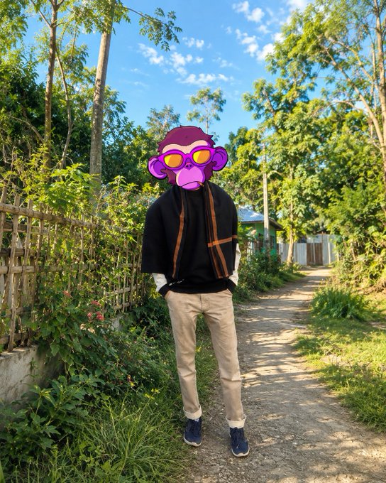 A photograph of a person standing on a dirt path outdoors, wearing a black jacket, beige pants, and blue shoes, with their face obscured by a purple cartoon monkey sticker wearing sunglasses.