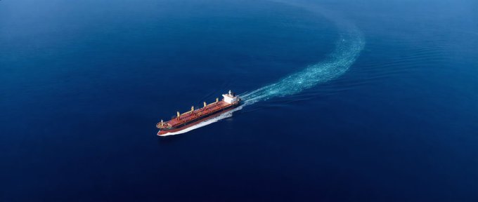 Aerial view of a red cargo ship sailing through deep blue ocean waters, leaving a curved wake behind it.