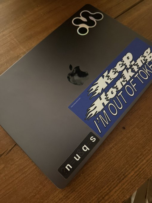 A silver Apple laptop rests on a wooden surface, decorated with stickers including one that reads "I'M OUT OF TOKENS" and another spelling "suan" with plastic letters.