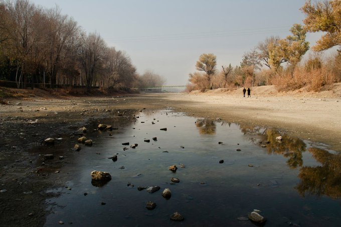 A serene landscape photograph of a shallow riverbed with scattered rocks, flanked by leafless trees and golden vegetation, with two small figures walking in the distance.