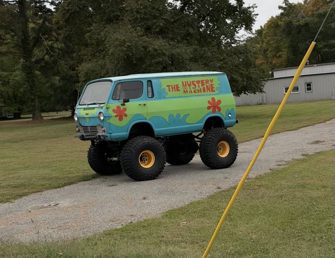 A lifted, vintage-style van painted in blue and green with flower decals is parked on a grass lawn; the side reads 'THE MYSTERY MACHINE'.