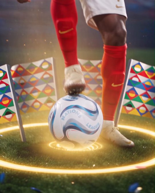 A close-up of a soccer player's foot wearing a red sock with a Nike swoosh and a white cleat resting on a 'NATIONS LEAGUE' soccer ball, surrounded by colorful geometric corner flags on a grass field with glowing yellow rings.