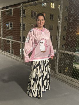 A woman standing in front of a chain-link fence wearing a pink sweater with a cartoon character design and a long patterned skirt.