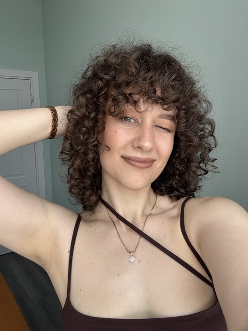 Selfie of a woman with curly brown hair winking at the camera, wearing a dark halter top and a delicate necklace with a small pendant.