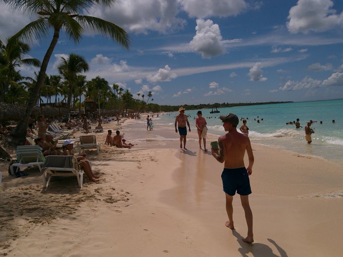 A sunny tropical beach scene with palm trees, lounge chairs, and people enjoying the turquoise water.