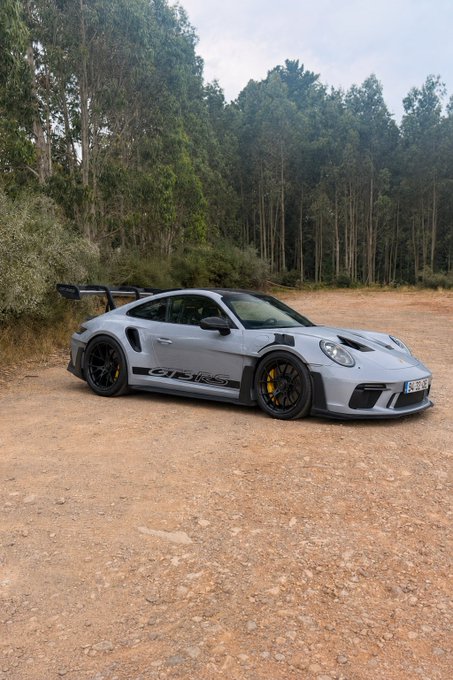 A silver Porsche 911 GT3 RS with yellow brake calipers and a prominent rear wing is parked on a dirt road lined with tall trees. The "GT3 RS" lettering is visible on the door, fitting the post's claim that crypto gains funded the high-performance sports car.