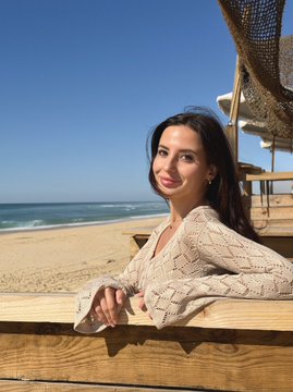 A young woman with dark hair smiles at the camera while leaning on a wooden railing, with a sandy beach and ocean visible behind her under a clear blue sky.