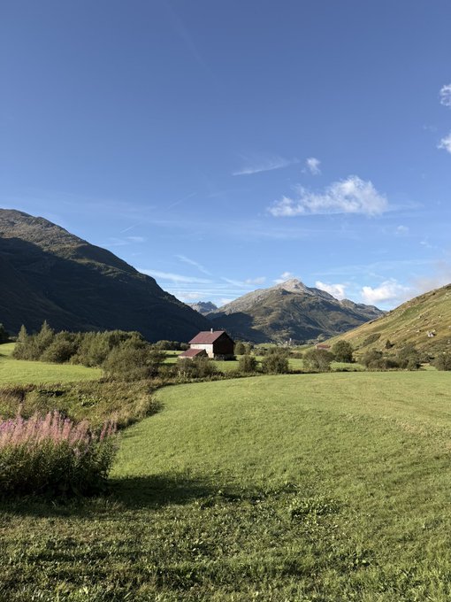 A lush green alpine meadow dotted with pink wildflowers stretches toward a solitary red barn nestled at the base of towering, rugged mountains under a bright blue sky. The serene landscape evokes the "GM" (Good Morning) greeting, offering a peaceful, scenic start to the day.